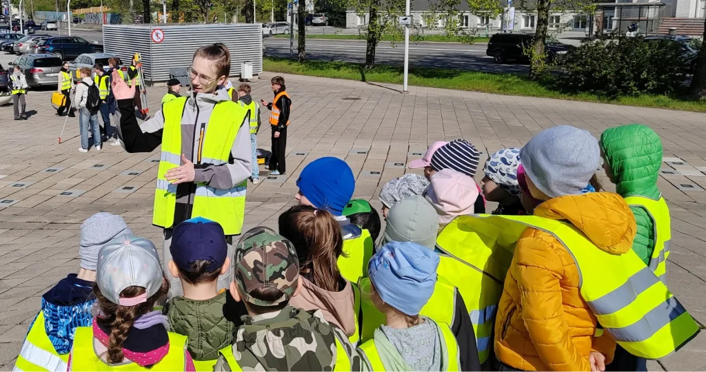 TTK kõrgkooli üliõpilane helkurlvestis räägib ülikooli esisel platsil lasteaialaste rühmale geodeetilistest mõõtmistöödest. Lapsed kuulavad huviga, taustal on näha teisi tudengeid ja õppevahendeid.